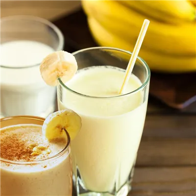3 glasses filled with milky drink with straws in them, on a wooden table with a bunch of bananas in the background.