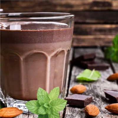 Chocolate Milk drink in a clear glass, on a wooden table scattered with pieces of dark chocolate, mint leaves and almonds.