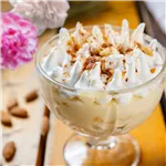 Almond Pudding in a glass dessert cup, topped with cream and crushed almonds, on a wooden table.