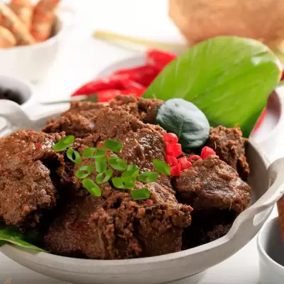 Meat rendang in white bowl garnished with chives, chillie and tumeric leaf.