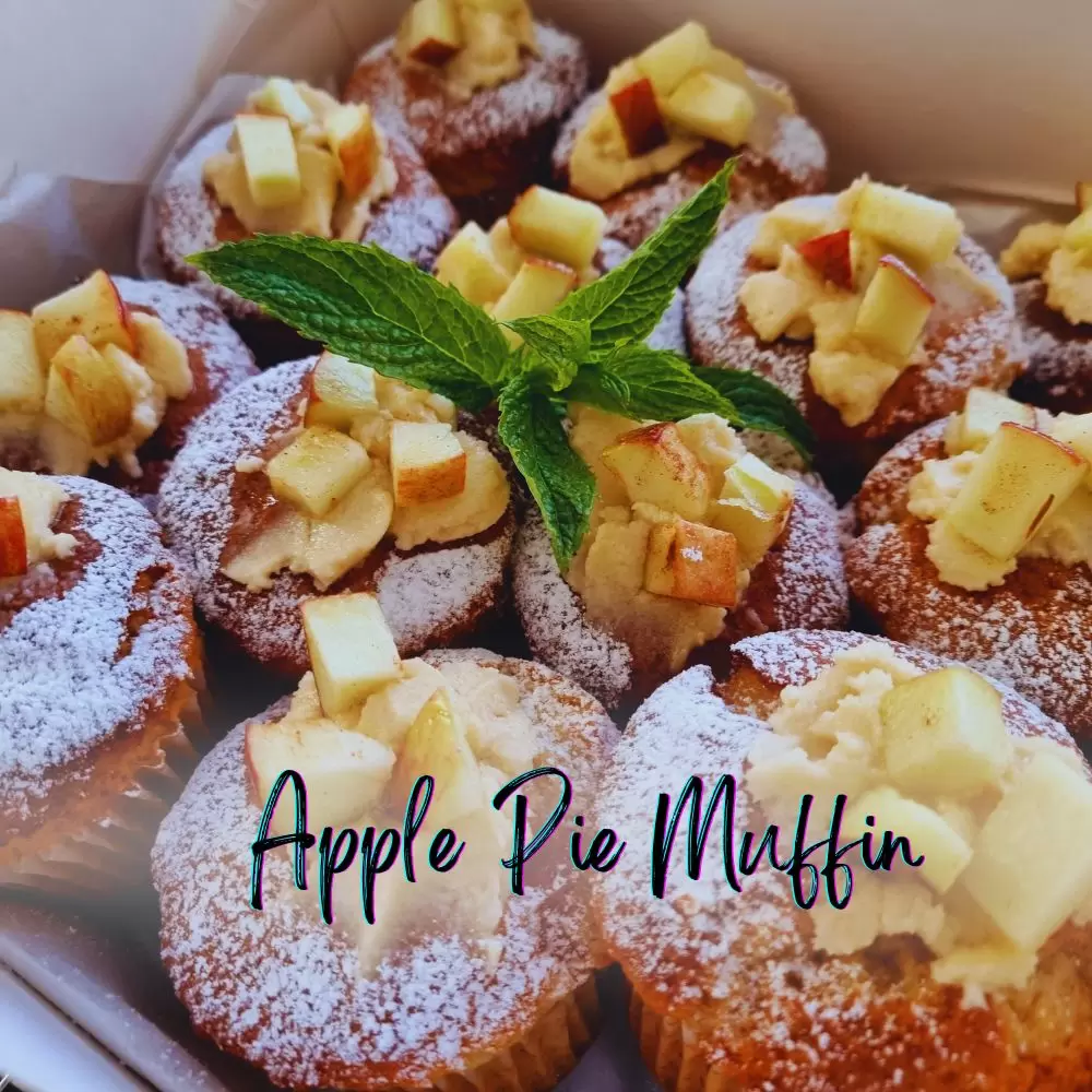 A box of apple pie muffins dusted with icing sugar and topped with diced apple and mint
