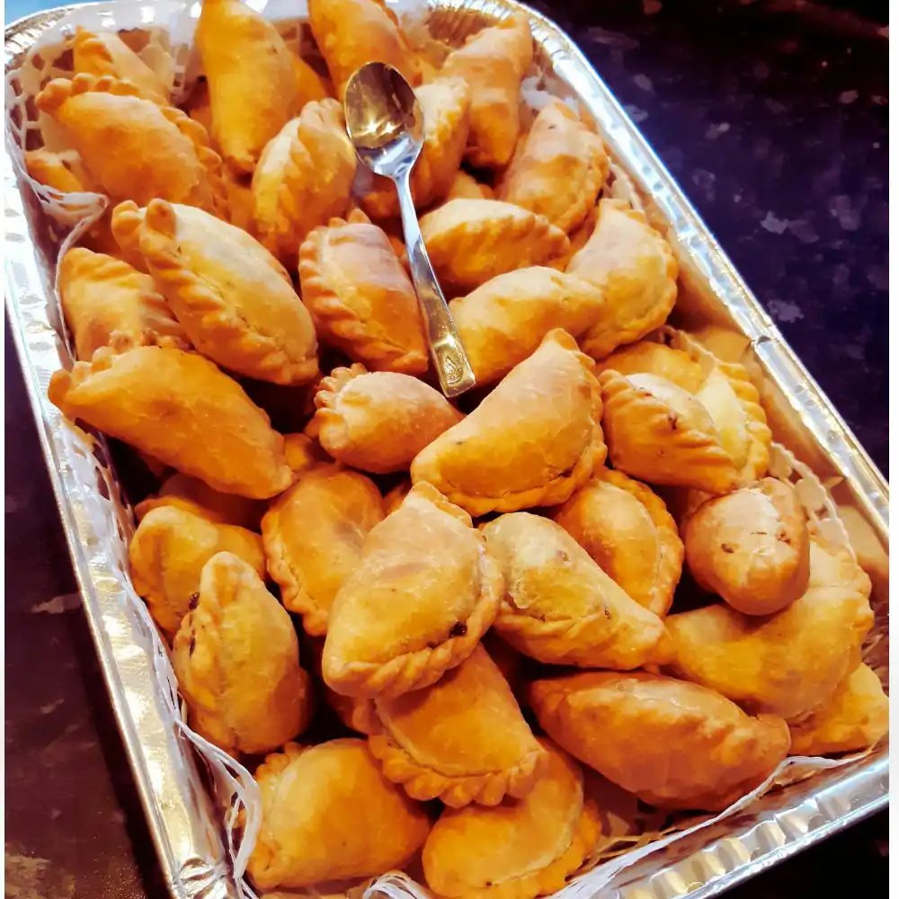 A foil catering tray piled high with golden baked pizza puff pastries and a serving spoon